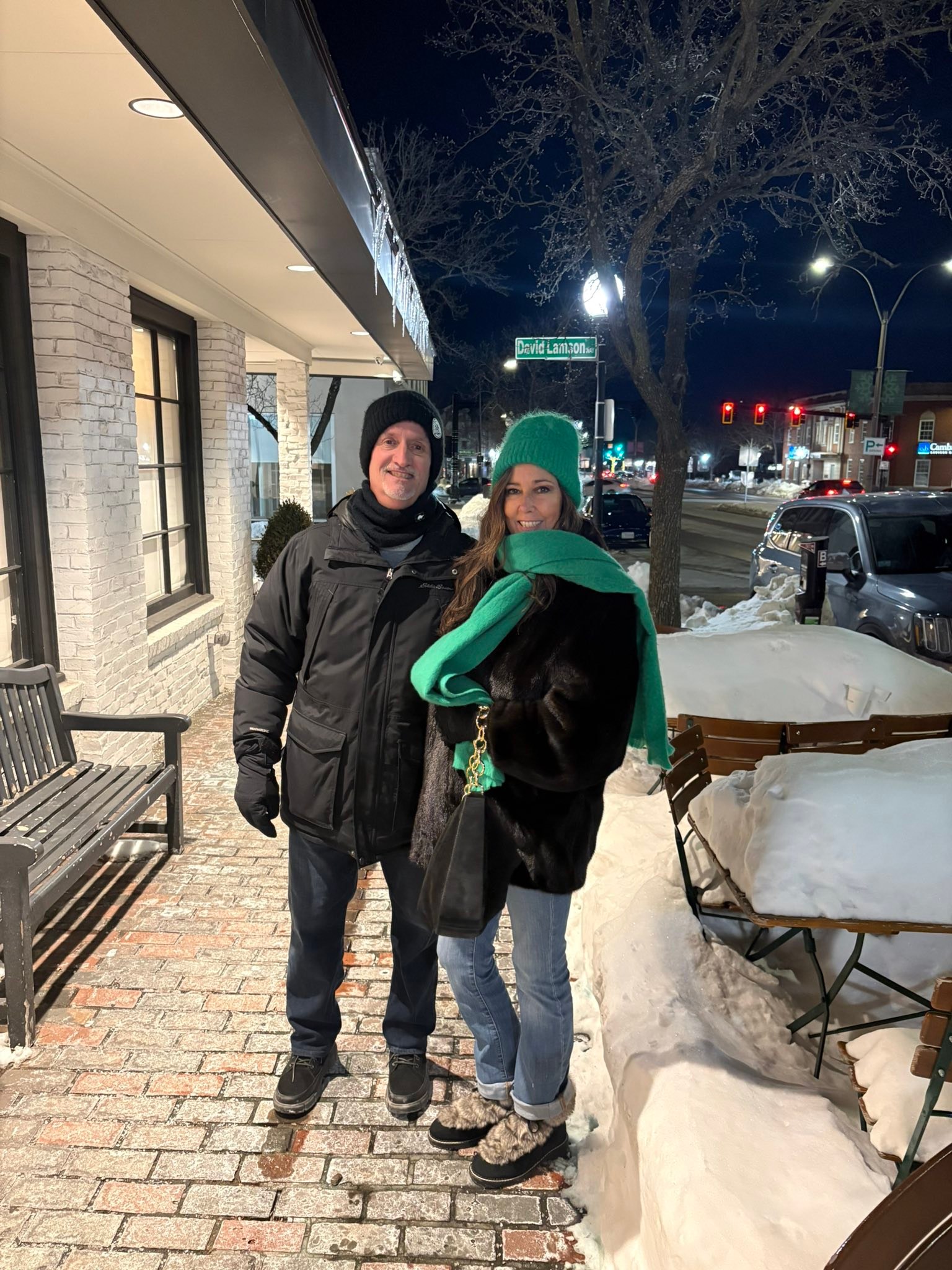 Two adults in winter clothing stand together outside a brick storefront on a snowy sidewalk at night, smiling at the camera (woman in green hat and scarf).