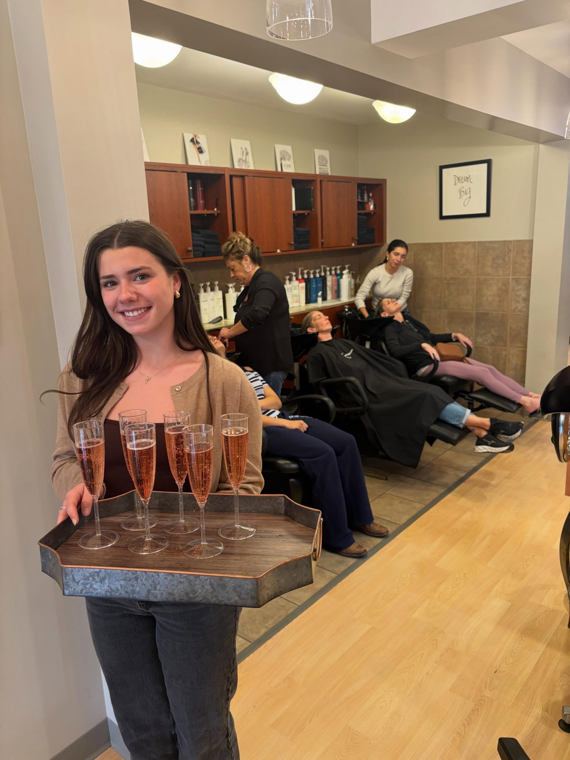 Smiling young woman holds a tray of six champagne flutes in a hair salon, with clients getting hair treatments in the background.