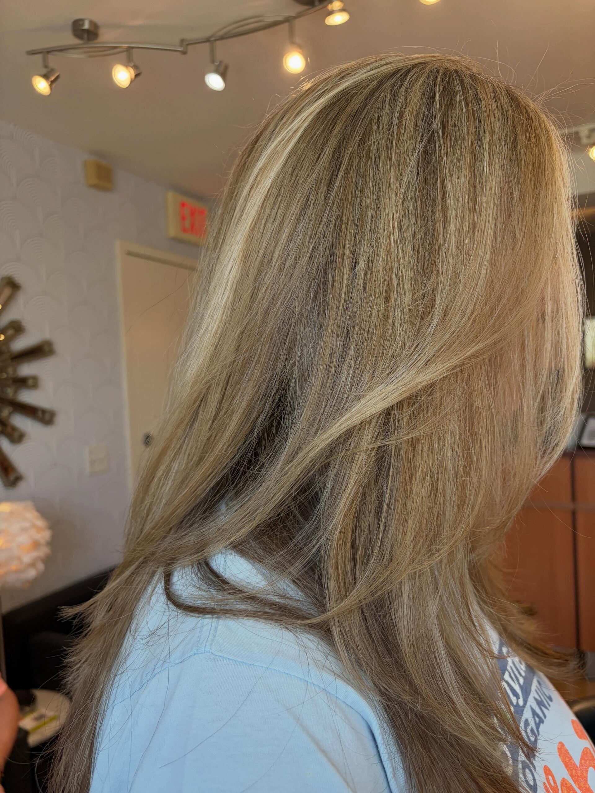 Back of a person’s head with long blonde hair, wearing a light blue shirt, in a room with track lighting and decorative walls in the background.