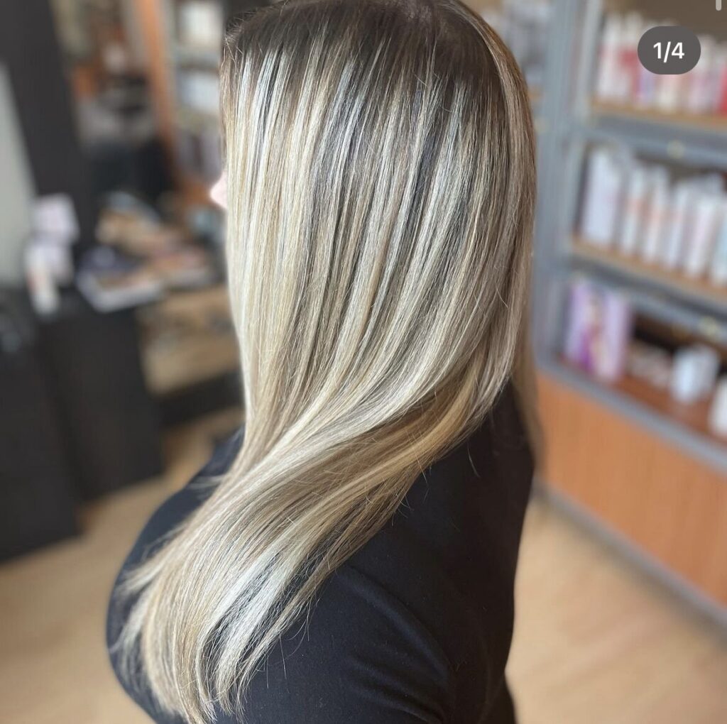 Woman with straight, shoulder-length blonde hair in a salon, showing warm caramel and cool platinum highlights from the side.