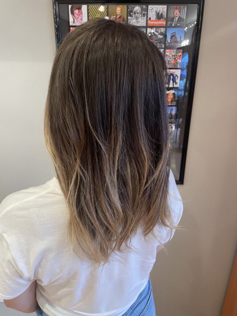 Back view of a person with medium-length brown hair with blonde ends, wearing a white t-shirt and blue jeans, standing in front of a collage photo frame on the wall.