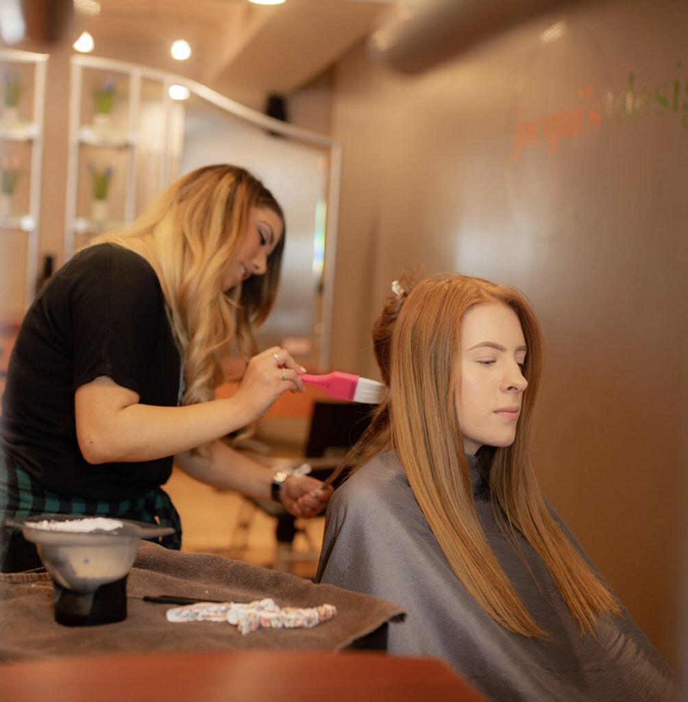 Hair stylist brushing the long blonde hair of a seated client in a salon.