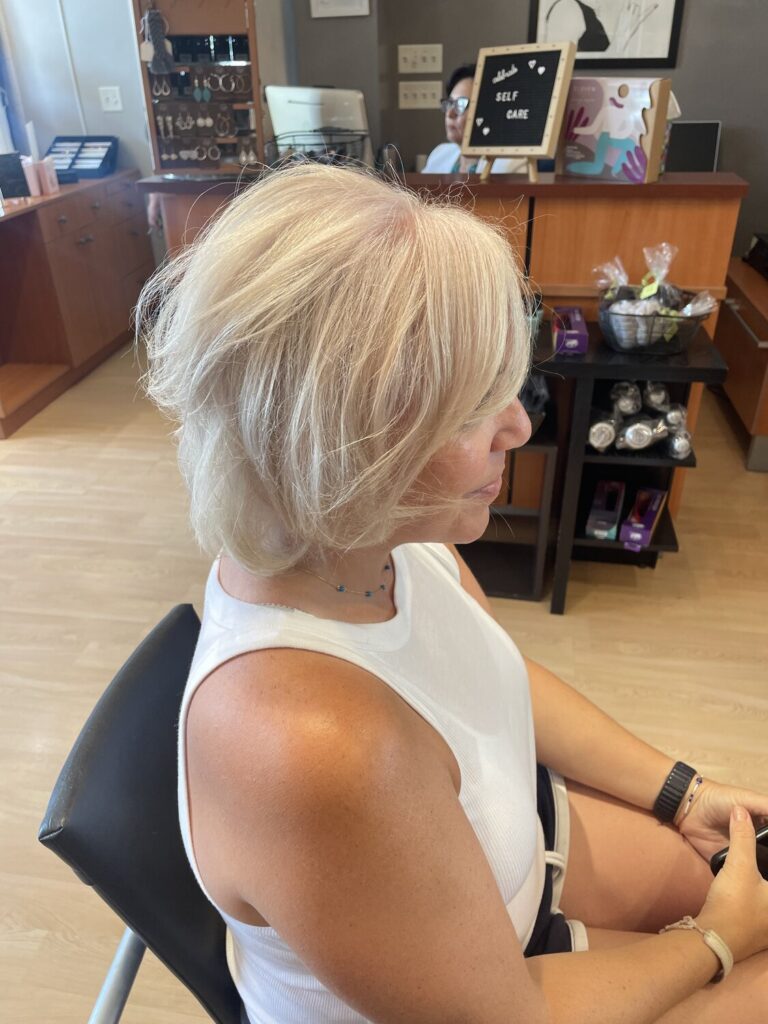 Side profile of a woman with short blonde hair seated in a salon chair, white tank top, in a salon with mirrors and product shelves behind her.