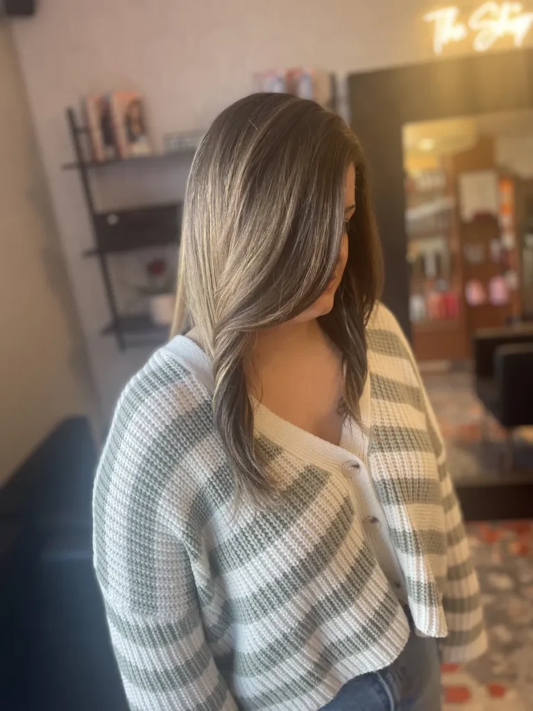 Woman in a green-and-white plaid cardigan with shoulder-length silver-blonde hair styled in loose waves, standing in a salon.