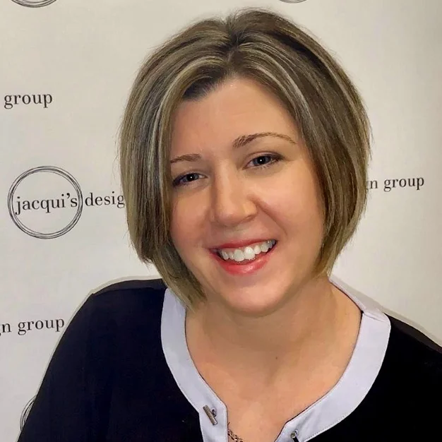 Smiling woman with a short brunette bob with blonde highlights, wearing a black top with a light gray collar, posing in front of a backdrop that reads 'jacqui's design group'