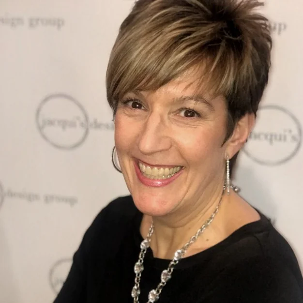 Smiling woman with short brown hair wearing a black top and silver jewelry, posing for a photo at an event backdrop.
