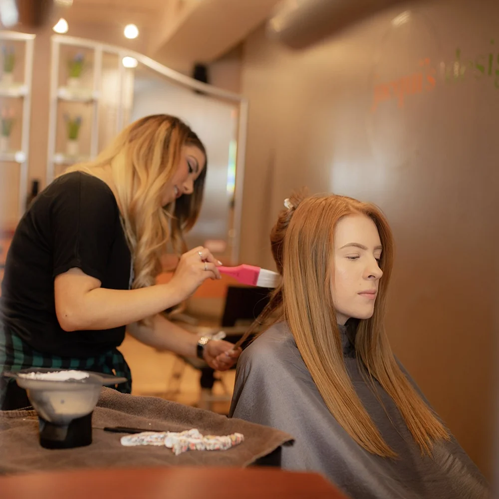 Hair stylist brushes a client's long blonde hair in a salon, cape on, focused on styling.
