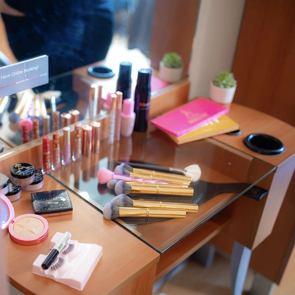 A makeup vanity with brushes, lipsticks, powders, and a mirror on a wooden desk, plus small potted plants in the background.