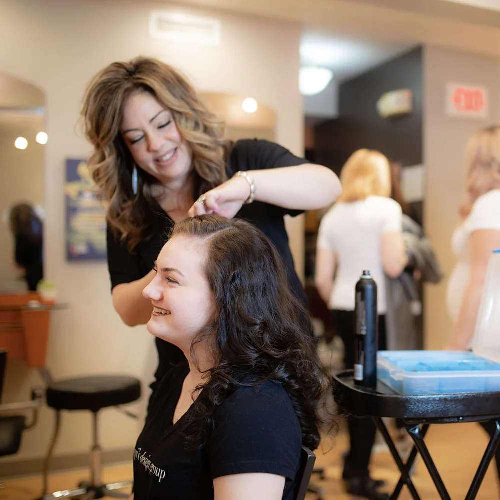 Hairdresser styles a smiling woman's hair in a busy salon, with styling cart and tools nearby.