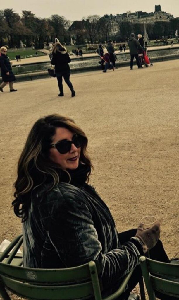 Woman with dark hair and sunglasses sits on a green park bench, holding a cup and smiling at the camera, with a fountain and pedestrians in the background.