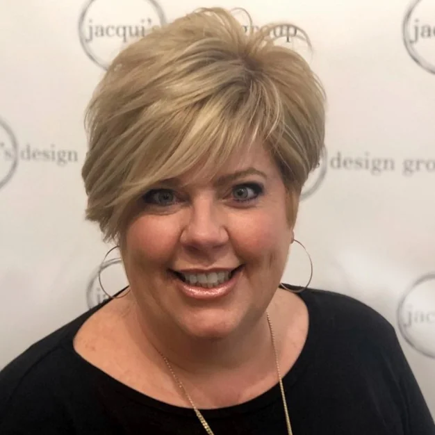 Portrait of a smiling woman with short blonde hair wearing a black top, in front of a Jacqu Design Group backdrop.