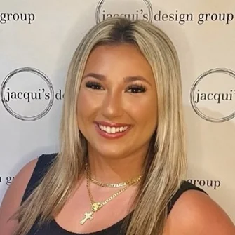 Smiling woman with shoulder-length blonde hair wearing a black top and gold necklaces, standing in front of a backdrop with jacqui's design group logos.