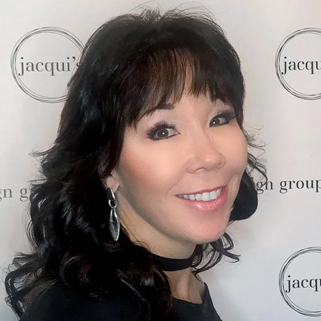 Smiling woman with dark, styled hair and hoop earrings posing at an event backdrop with jacqui's logo; wearing a black choker and subtle makeup.