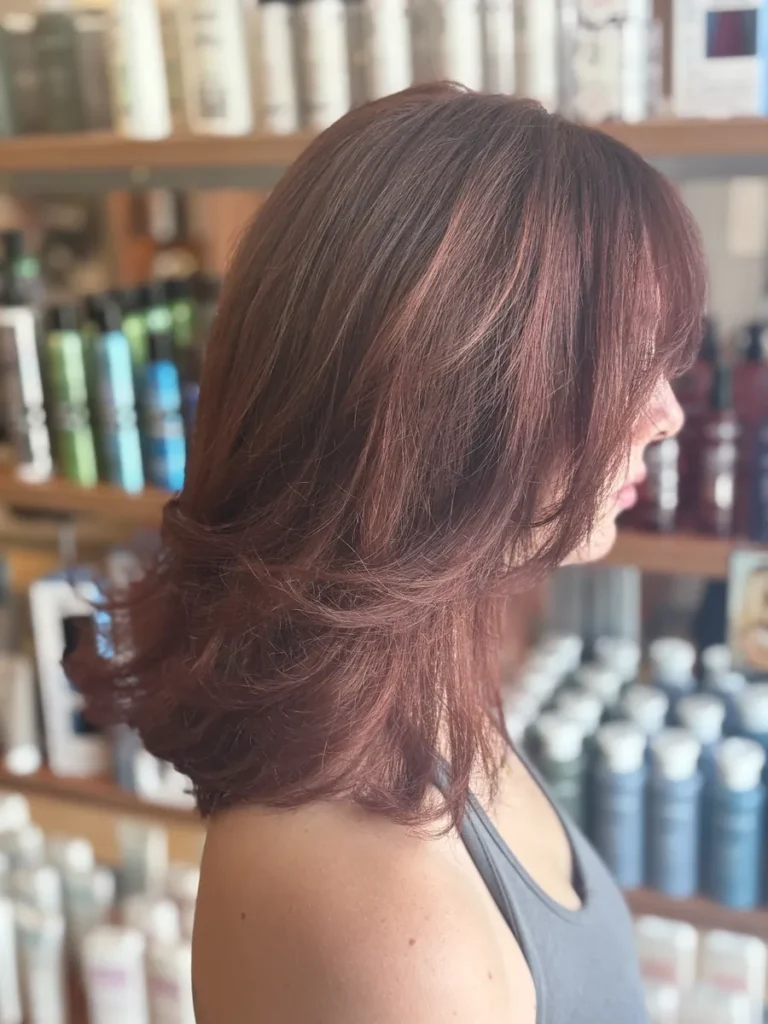 Side view of a woman with layered, shoulder-length brown hair with reddish highlights in a salon, shelves of hair products in the background.