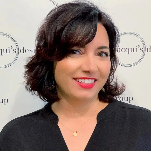 Portrait of a smiling woman with short dark hair wearing a black blouse, standing in front of a branded backdrop.