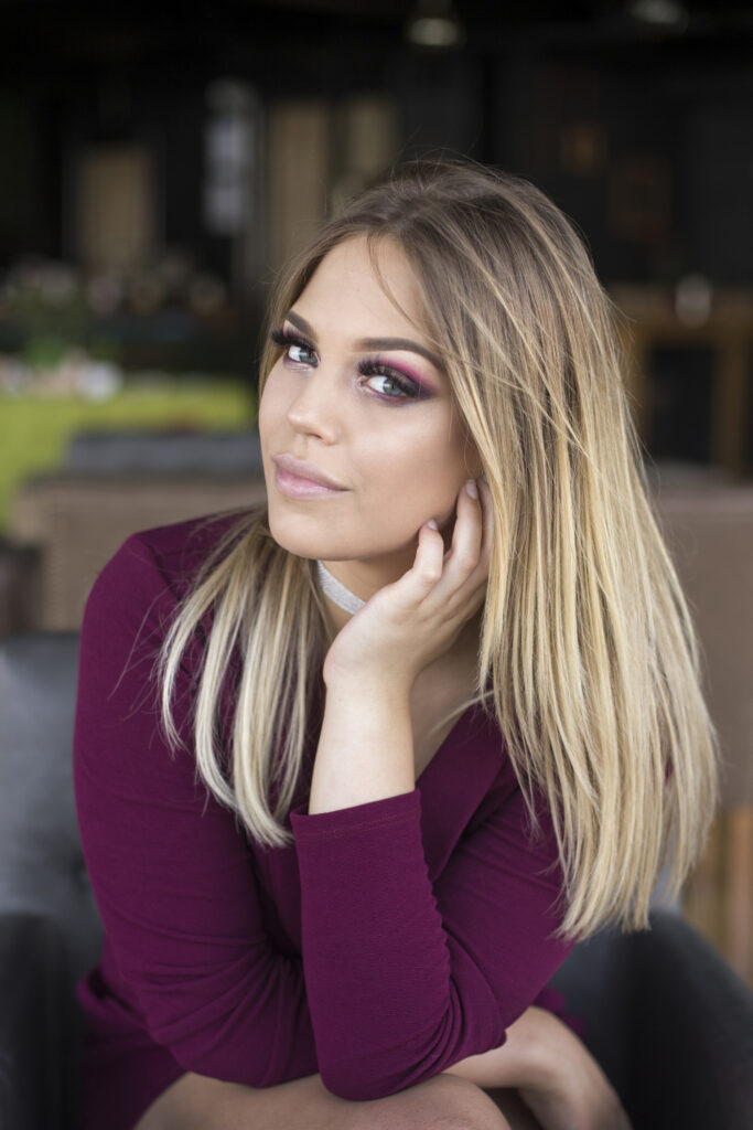 Portrait of a young woman with long blonde hair and pink makeup resting her chin on her hand in a blurred indoor background.