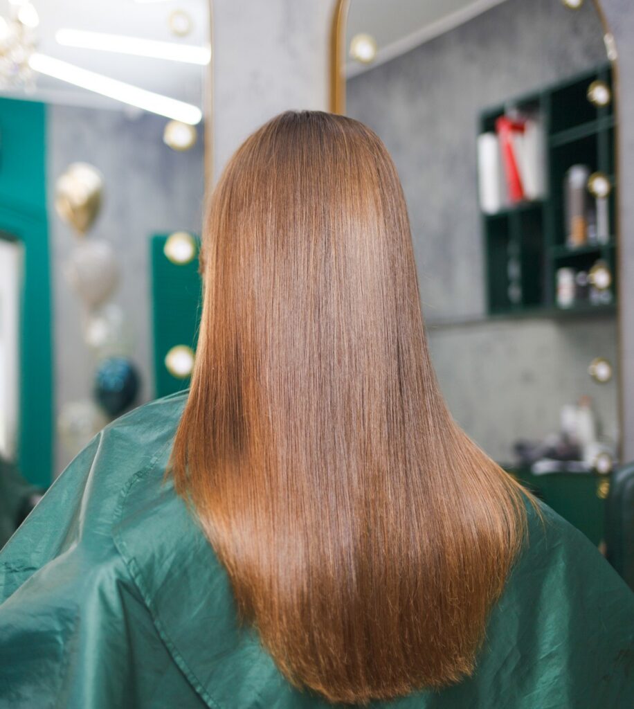 Person with long straight brown hair seen from behind in a hair salon, wearing a green cape.